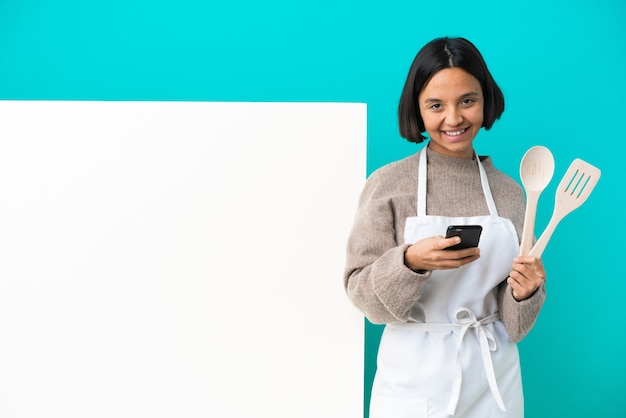 モバイルでメッセージを送信する青い背景で隔離の大きなプラカードを持つ若い混血料理人の女性