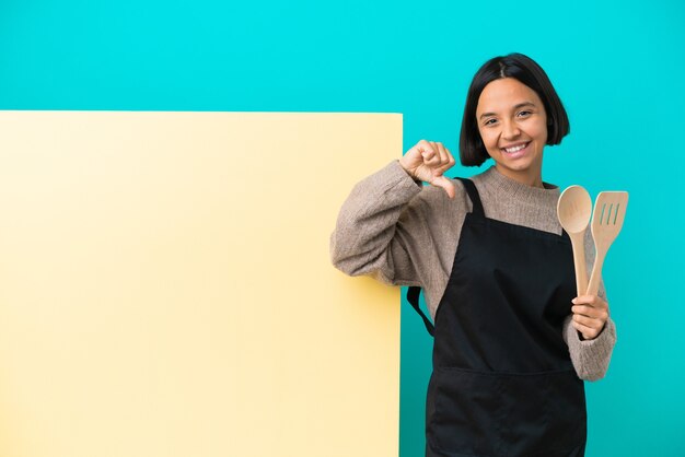 Young mixed race cook woman with a big placard isolated on blue background showing thumb down with negative expression