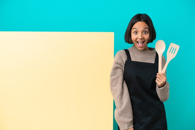 Young mixed race cook woman with a big placard isolated on blue background with surprise facial expression