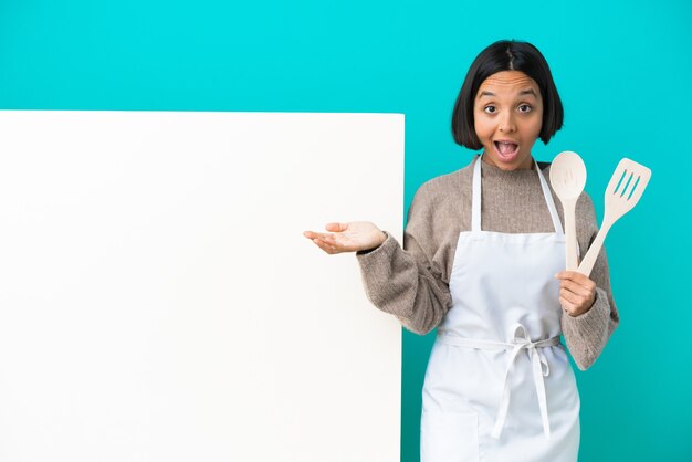 Young mixed race cook woman with a big placard isolated on blue background with surprise and shocked facial expression