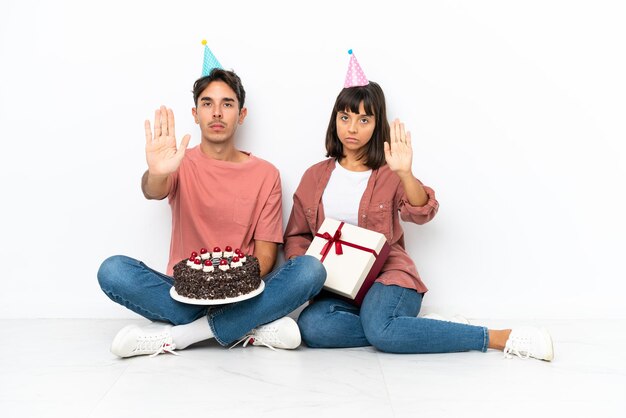 Young mixed race couple celebrating a birthday sitting on the floor isolated on white background making stop gesture denying a situation that thinks wrong