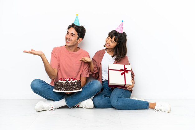 Молодая пара смешанной расы празднует день рождения, сидя на полу на белом фоне, указывая назад и представляя продукт