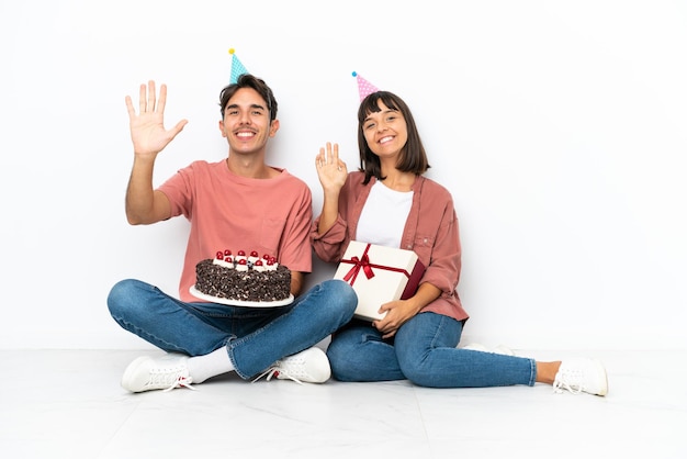 Young mixed race couple celebrating a birthday sitting on the floor isolated on white background saluting with hand with happy expression