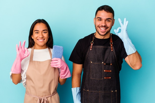 Young mixed race couple cleaning home isolated on blue background cheerful and confident showing ok gesture.