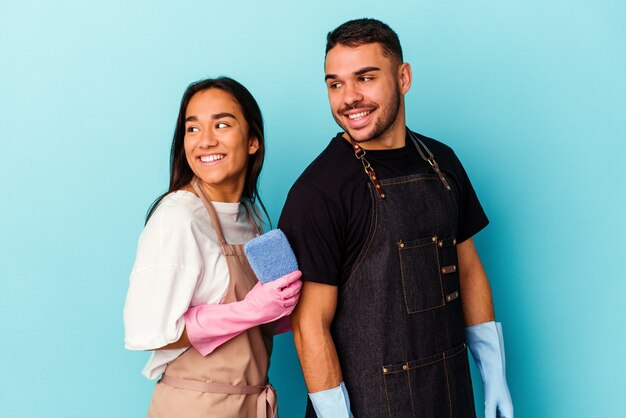 Young mixed race couple cleaning home isolated on blue looks aside smiling, cheerful and pleasant.