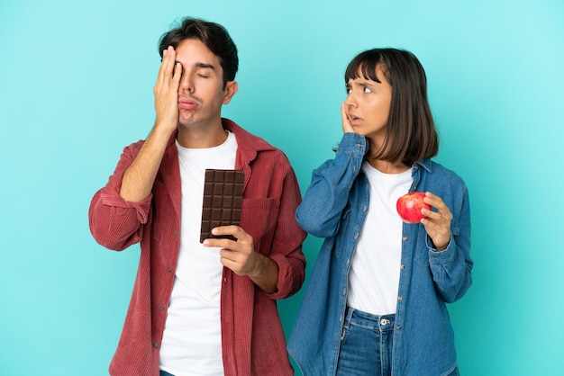 Молодая пара смешанной расы, держащая яблоко и шоколад на синем фоне с удивленным и шокированным выражением лица