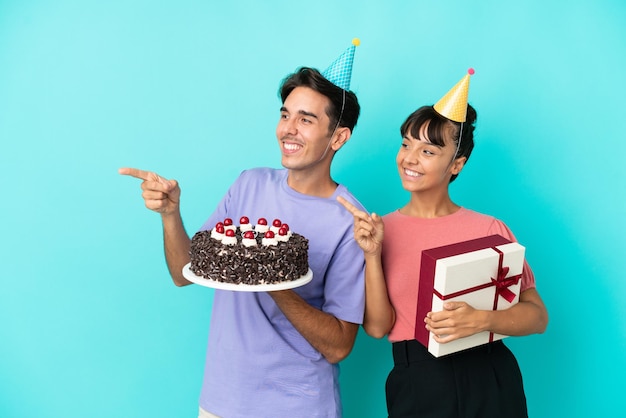 Giovane coppia di razza mista in possesso di torta di compleanno e presente isolato su sfondo blu che punta il dito di lato in posizione laterale