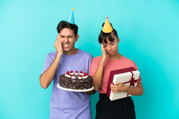 Young mixed race couple holding birthday cake and present isolated on blue background unhappy and frustrated with something