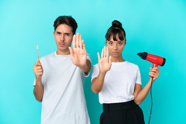 Young mixed race couple holding a hairdryer and toothbrush\
isolated on blue background making stop gesture denying a situation\
that thinks wrong