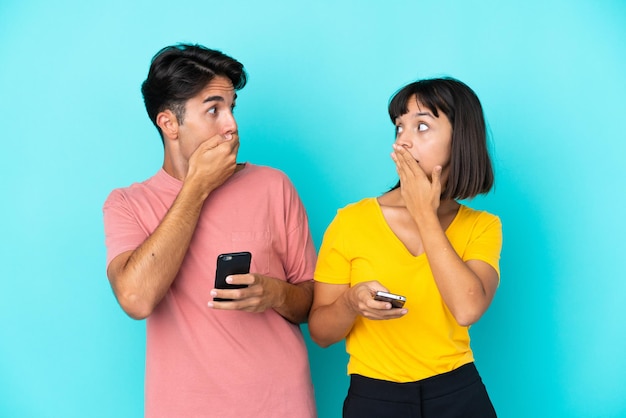 Photo young mixed race couple holding mobile phone isolated on blue background covering mouth with hands for saying something inappropriate