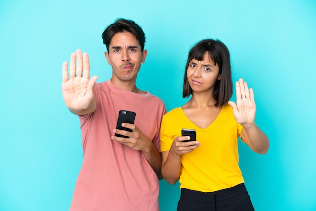 Young mixed race couple holding mobile phone isolated on blue background making stop gesture denying a situation that thinks wrong