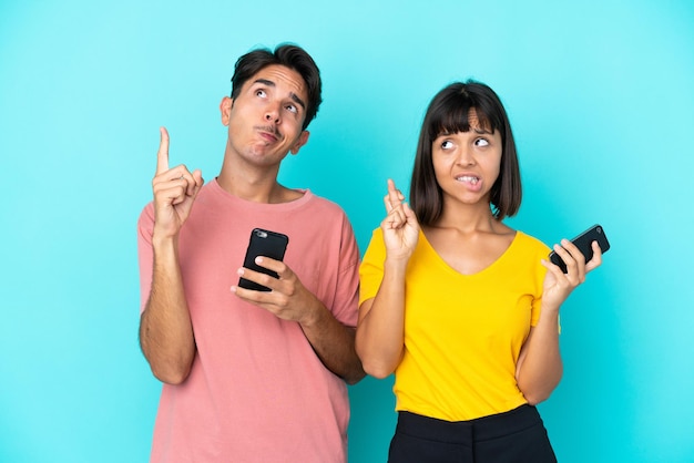 Young mixed race couple holding mobile phone isolated on blue background with fingers crossing and wishing the best