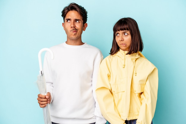 Young mixed race couple holding umbrella isolated on blue background confused, feels doubtful and unsure.