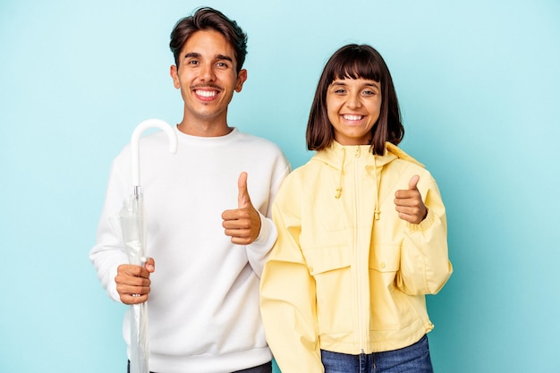 young mixed race couple holding umbrella isolated on blue background smiling and raising thumb up