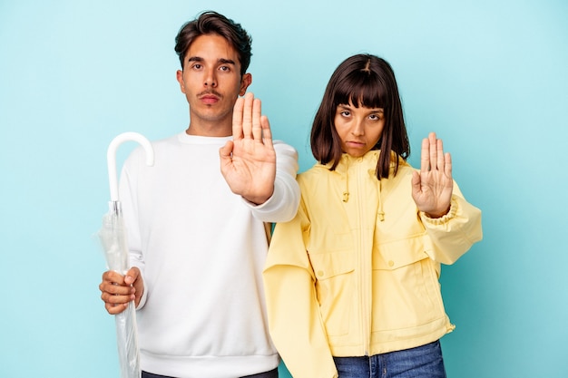 Young mixed race couple holding umbrella isolated on blue background standing with outstretched hand showing stop sign, preventing you.