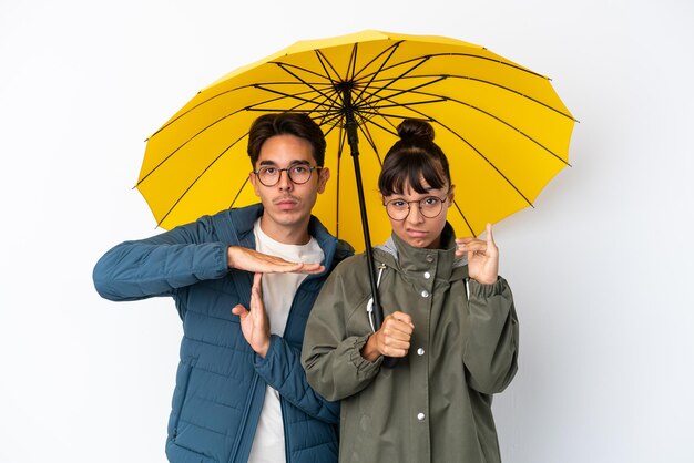 Young mixed race couple holding an umbrella isolated on white background making stop gesture with her hand to stop an act