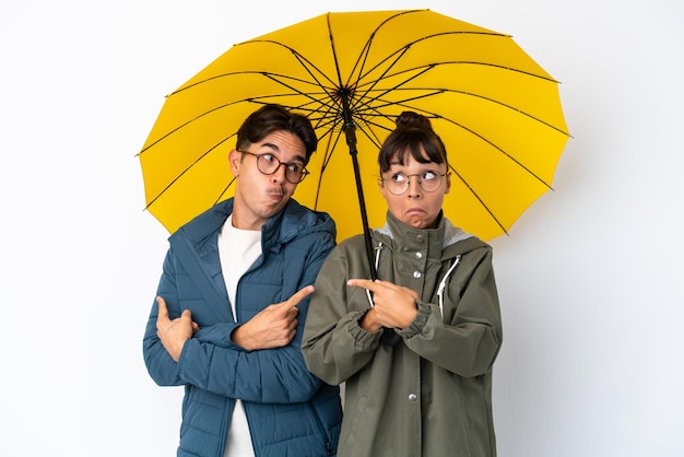 Young mixed race couple holding an umbrella isolated on white background pointing to the laterals having doubts