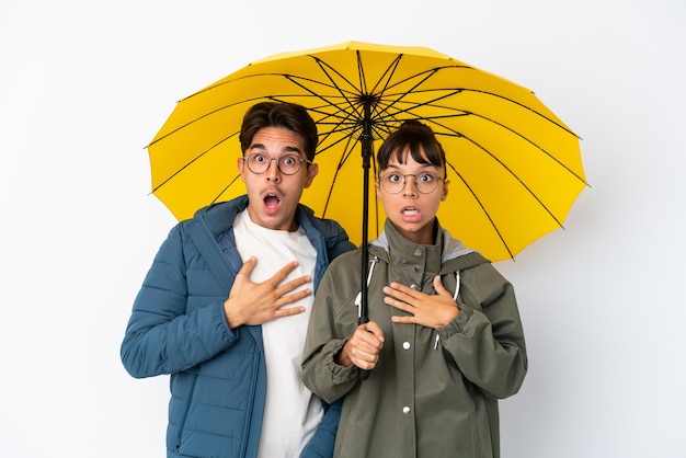 Young mixed race couple holding an umbrella isolated on white background surprised and shocked while looking right