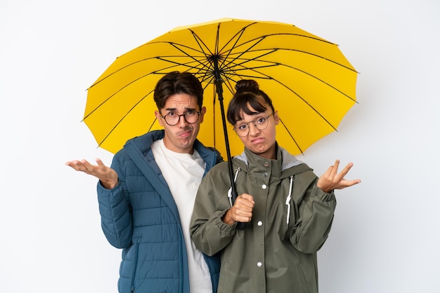 Young mixed race couple holding an umbrella isolated on white background unhappy and frustrated with something because not understand something