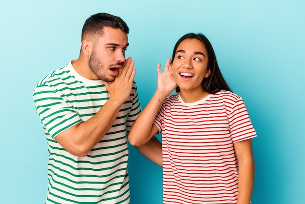 Young mixed race couple isolated on blue background trying to listening a gossip.