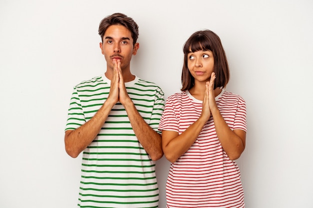 Young mixed race couple isolated on white background praying, showing devotion, religious person looking for divine inspiration.