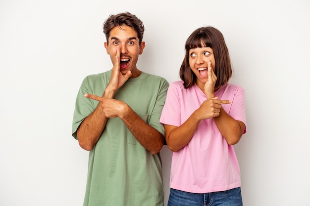 Young mixed race couple isolated on white background saying a gossip, pointing to side reporting something.