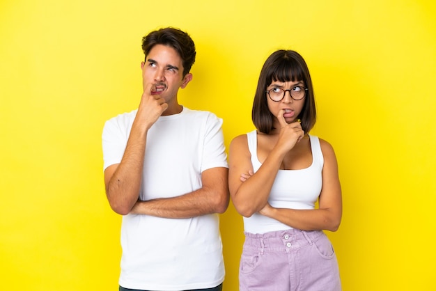 Young mixed race couple isolated on yellow background having doubts while looking up