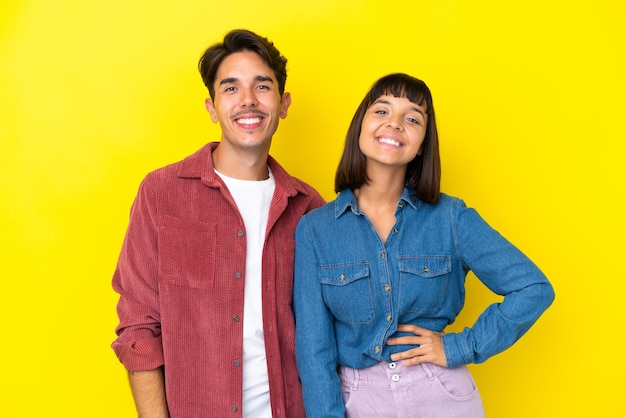 Young mixed race couple isolated on yellow background posing with arms at hip and smiling