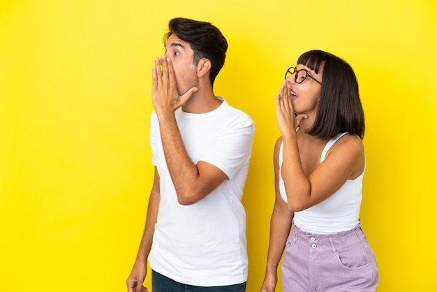 Photo young mixed race couple isolated on yellow background shouting with mouth wide open to the lateral