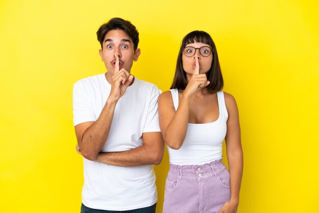 Young mixed race couple isolated on yellow background showing a sign of silence gesture putting finger in mouth