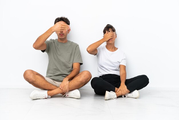 Young mixed race couple sitting on the floor isolated on white background covering eyes by hands Do not want to see something