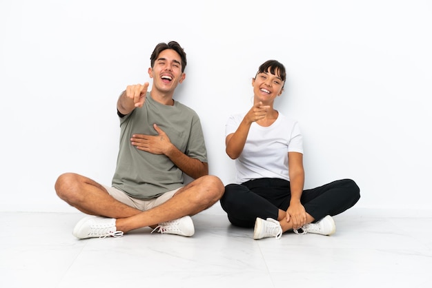 Молодая пара смешанной расы сидит на полу на белом фоне, указывая пальцем на кого-то и много смеясь