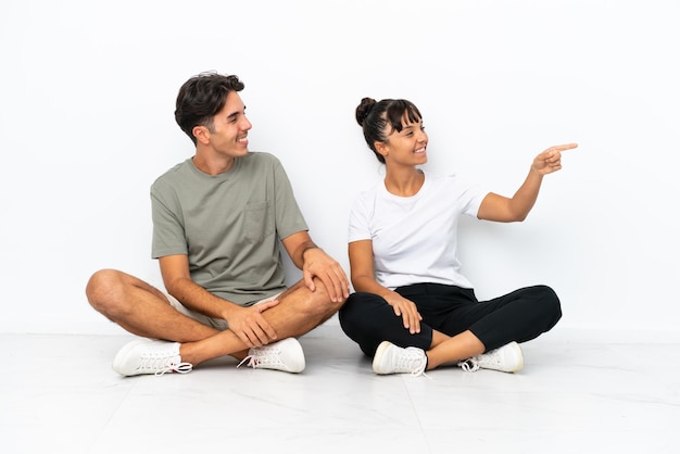 Young mixed race couple sitting on the floor isolated on white background presenting an idea while looking smiling towards