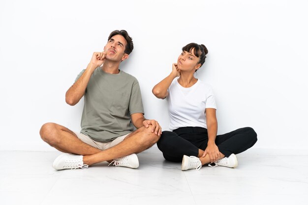 Young mixed race couple sitting on the floor isolated on white background thinking an idea while scratching head