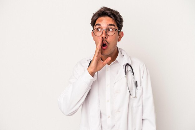 Young mixed race doctor man isolated on white background is saying a secret hot braking news and looking aside