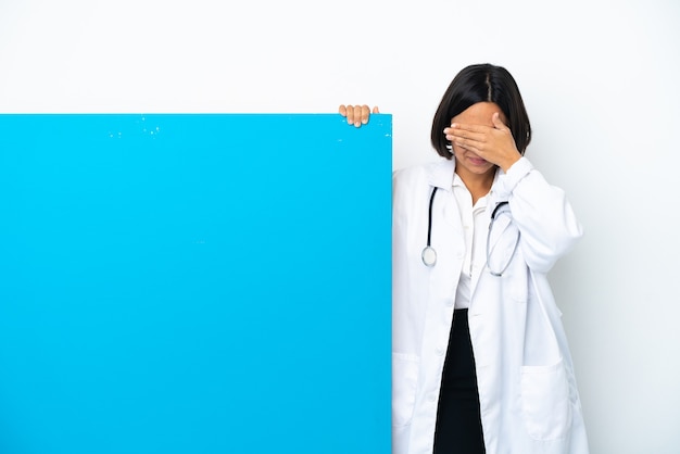 Young mixed race doctor woman with a big placard isolated on white background covering eyes by hands