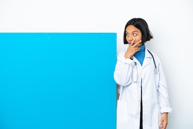 Young mixed race doctor woman with a big placard isolated on white background covering mouth and looking to the side