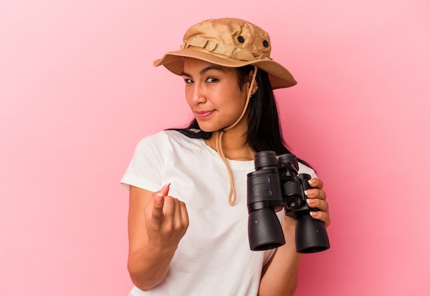 Young mixed race explorer woman holding binoculars isolated on pink background pointing with finger at you as if inviting come closer.