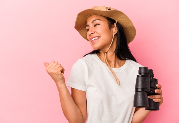 Young mixed race explorer woman holding binoculars isolated on pink background points with thumb finger away, laughing and carefree.