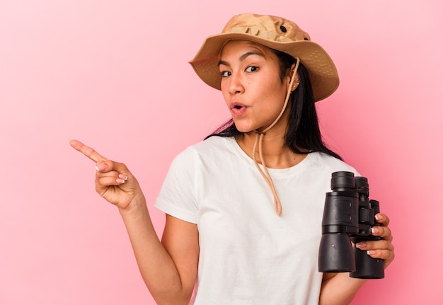 Young mixed race explorer woman holding binoculars isolated on pink wall pointing to the side