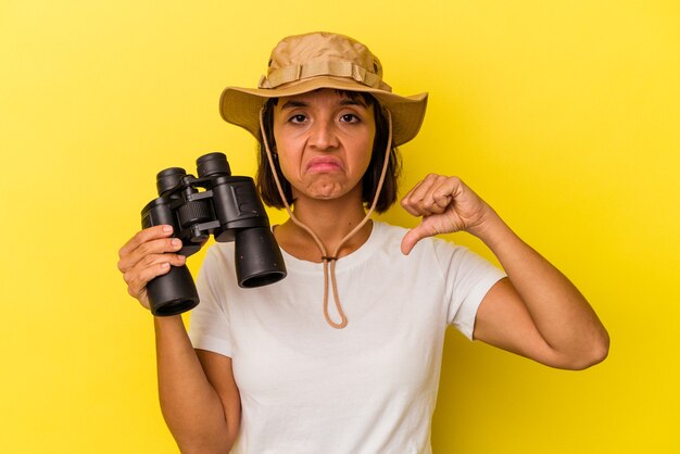 Young mixed race explorer woman holding binoculars isolated on yellow background showing a dislike gesture, thumbs down. Disagreement concept.
