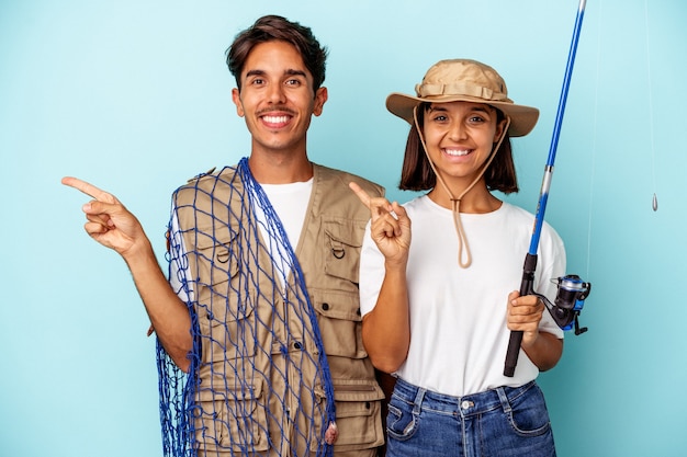 Young mixed race fisher couple isolated on blue background smiling and pointing aside, showing something at blank space.