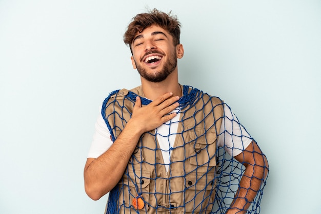 Young mixed race fisherman holding a net isolated on blue background laughs out loudly keeping hand on chest.