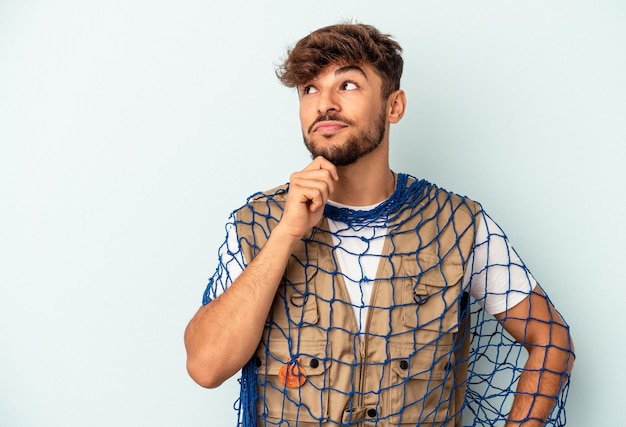 Young mixed race fisherman holding a net isolated on blue background looking sideways with doubtful and skeptical expression.