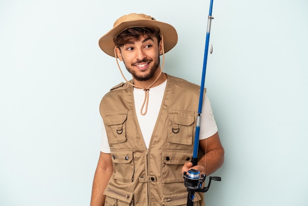 Young mixed race fisherman holding a rod isolated on blue background looks aside smiling, cheerful and pleasant.