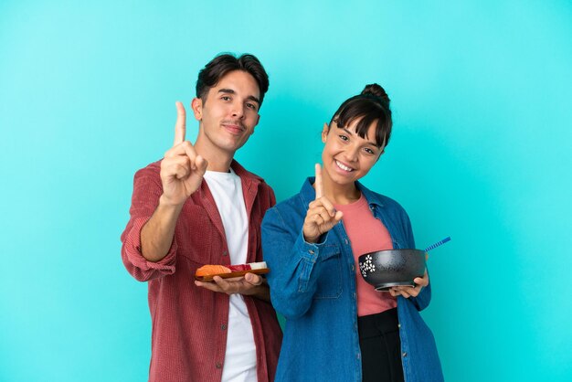 Young mixed race friends holding sashimi and ramen isolated on blue background showing and lifting a finger