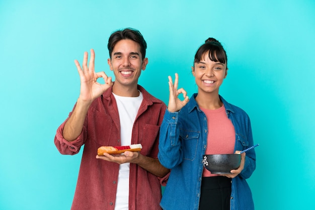 Young mixed race friends holding sashimi and ramen isolated on blue background showing an ok sign with fingers