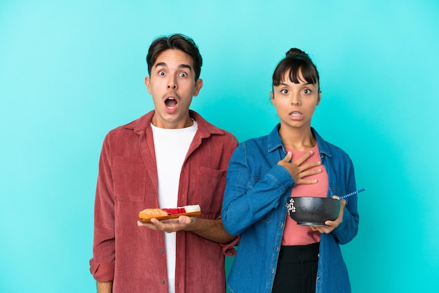 Young mixed race friends holding sashimi and ramen isolated on blue background with surprise and shocked facial expression