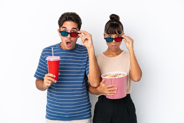 Giovani amici di razza mista isolati su sfondo bianco sorpresi con occhiali 3d e tenendo un grande secchio di popcorn