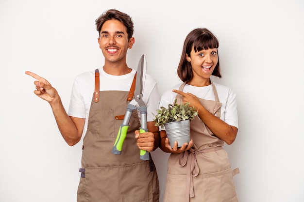 Young mixed race gardener couple isolated on white background smiling and pointing aside, showing something at blank space.